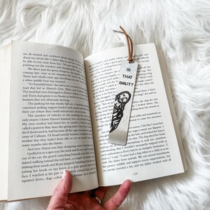 May include: A silver bookmark with a black and white illustration of a person with the text "Is That Smut?" printed on it.