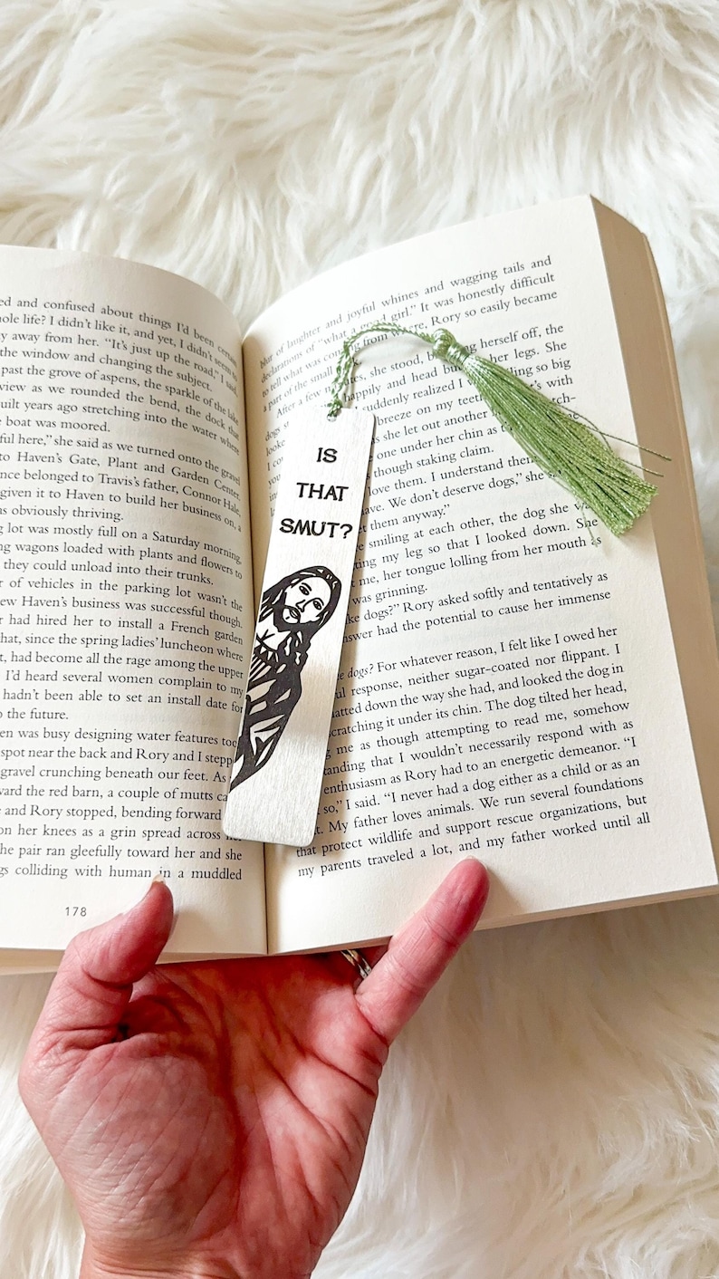 May include: A wooden bookmark with a green tassel. The bookmark features a black and white illustration of a man with a beard and the text "Is That Smut?" printed on it.