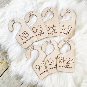 May include: Set of six wooden hangers with engraved numbers and text indicating age ranges in months for newborn to 24 months. The hangers are made of natural wood and have a simple, minimalist design.