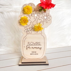 May include: Wooden craft with a vase and flowers. The vase is a mason jar with the text "Flowers for mommy" and "Picked by Hunter & Hudson". The flowers are yellow and red.
