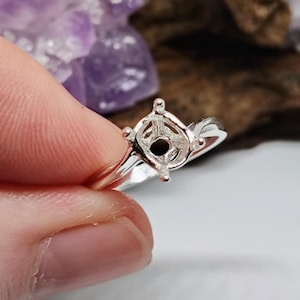 May include: A silver ring with a prong setting, designed to hold a gemstone. The ring is held in a hand, with a blurred background of purple crystals and wood.