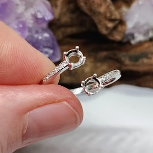 May include: A silver-toned open ring setting featuring two circular prongs designed to hold gemstones. The band is accented with small, clear pavé-set stones along the sides, creating a delicate and refined appearance for custom jewellery assembly.