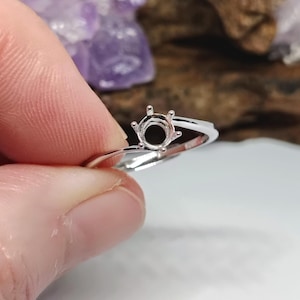 May include: A silver ring with a round setting, held by a hand. The ring's setting is designed to hold a gemstone. The ring is on a white surface, with a purple crystal and wood in the background.