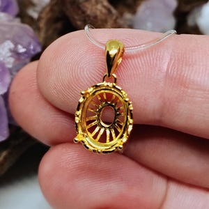 May include: A gold pendant with an oval setting. The pendant features an openwork design and is suspended from a clear cord. The background includes natural elements, such as wood and crystals. The pendant is likely a piece of jewellery.