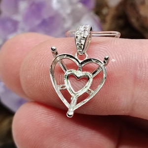 May include: A silver pendant with a heart-shaped design. The pendant features a smaller heart inside a larger heart, with a decorative bail adorned with small stones. The pendant is held between fingers.