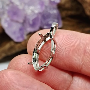 May include: A silver-colored pendant setting with an oval shape, featuring prongs for securing a gemstone. The setting has a loop at the top for a chain. The background includes a blurred purple crystal and wood.