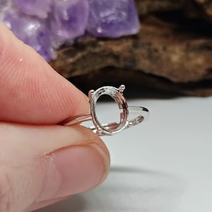 May include: A silver ring setting with an oval-shaped bezel. The ring is held by a hand, with a blurred background featuring a purple crystal and wood.