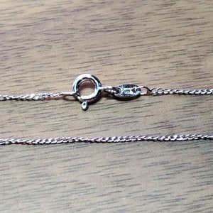 Op de afbeelding: Een zilverkleurige ketting met een karabijnsluiting. De ketting is gemaakt van kleine, strak verbonden segmenten, waardoor een getextureerd uiterlijk ontstaat. De sluiting is rond met een kleine hendel om te openen en te sluiten. De ketting wordt gepresenteerd op een houten oppervlak.