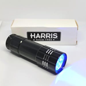 Op de afbeelding: Een zwarte zaklamp met blauw licht dat van voren komt. De zaklamp heeft witte strepen en de tekst "HARRIS Since 1922" is zichtbaar op de witte doos erachter.