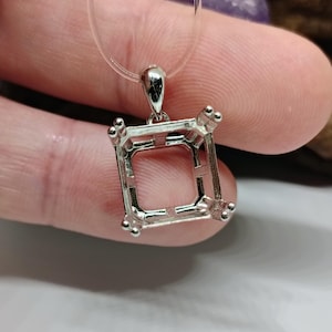 Puede incluir: Un colgante plateado con un engaste cuadrado, diseñado para sostener una piedra preciosa. El colgante está suspendido de un cordón transparente. El engaste tiene cuatro garras en cada esquina para asegurar la piedra.