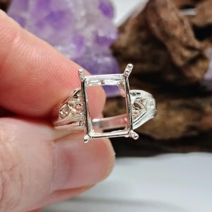 May include: A silver ring with an open rectangular setting, designed to hold a gemstone. The ring features intricate filigree detailing on the band and around the setting. The ring is held in a hand, with a blurred background of purple and brown.