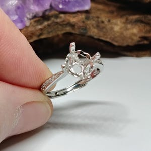 Puede incluir: Un anillo plateado con engaste de garras, diseñado para sostener una piedra preciosa. La banda presenta pequeños detalles brillantes. El anillo es sostenido por una mano contra un fondo borroso.