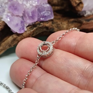 May include: A delicate silver bracelet featuring a circular pendant encrusted with small, sparkling stones. The bracelet's chain is composed of small, interconnected links. The background includes a cluster of purple crystals and a piece of wood.