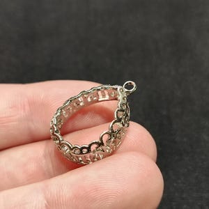 Puede incluir: Un colgante plateado en forma de lágrima calada con un pequeño lazo en la parte superior. El colgante presenta un delicado diseño floral o de volutas repetido en los bordes. El fondo es de color oscuro y liso.