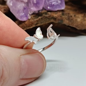 May include: A silver butterfly ring with white accents. The ring features two butterfly designs, one on each end of the adjustable band. The ring is held by a person's fingers, with a blurred background of purple crystals and wood.