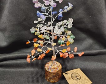 May include: A handcrafted wire tree sculpture with colorful gemstone leaves. The tree features a trunk made of a small wooden cylinder filled with stones. The gemstones are in shades of purple, blue, green, orange, and clear. A small tag is attached.