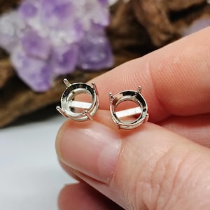 May include: Two silver-colored, round earring settings with four prongs each, designed to hold gemstones. The settings have a horizontal bar across the center. The background includes a blurred purple crystal and wood.