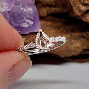 May include: A silver ring with a pear-shaped setting, designed to hold a gemstone. The band features a delicate, textured design with small, sparkling accents. The ring is held against a blurred background with purple and brown tones.