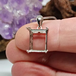 May include: A silver-colored pendant setting, rectangular in shape, with four prongs at the corners. The setting is held between fingers, with a blurred background of purple amethyst and brown wood.