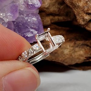 Puede incluir: Un engaste de anillo plateado con un centro rectangular y una banda adornada con pequeñas piedras brillantes. El anillo se sostiene contra un fondo borroso de elementos morados y marrones.