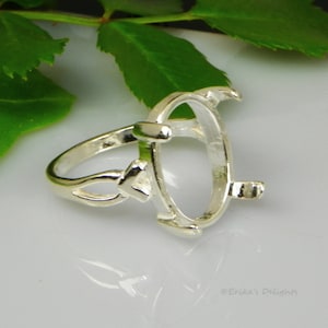 May include: Silver ring setting with an oval shaped bezel for a gemstone. The ring has a delicate design with a twisted band and a small decorative element on the side.