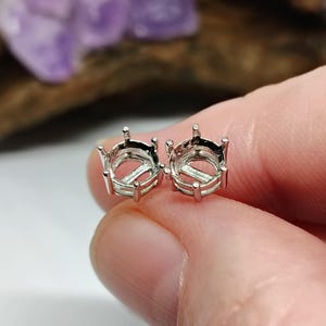 May include: Two silver-colored jewelry settings, held between fingers, are shown in a close-up shot. Each setting has a circular design with prongs extending upwards. The settings are designed to hold gemstones or other decorative elements.