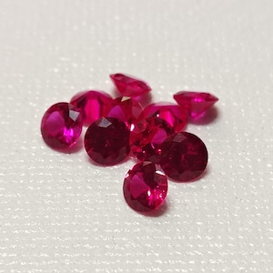 May include: A collection of faceted, round-cut rubies. The gemstones are a deep, vibrant red color, with multiple facets catching the light. The rubies are scattered on a white surface, showcasing their brilliance.