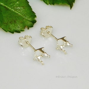May include: A pair of silver-colored earring settings, each with a post and a four-prong setting designed to hold a gemstone. The earrings are displayed on a white surface with a green leaf in the background.
