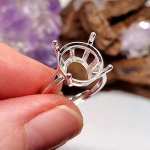 May include: A silver-colored ring setting with a round, open design to hold a gemstone. The ring has four prongs and an adjustable band. The setting is held up by a person's fingers, with a blurred background of wood and purple.