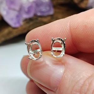 Puede incluir: Dos engastes de pendientes ovalados plateados. Los engastes se sostienen entre los dedos, con un fondo borroso de tonos morados y marrones. Los engastes están diseñados para sujetar gemas ovaladas.