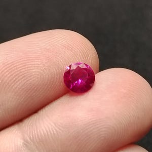 Peut inclure: Une pierre précieuse de rubis ronde et facettée. La gemme est d'une teinte rouge vibrante, avec de multiples facettes captant la lumière. Le rubis est tenu entre deux doigts sur un fond flou.