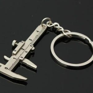 May include: Silver-toned metal keychain shaped like a caliper with a ruler marked in inches. The keychain has a silver-toned metal ring.