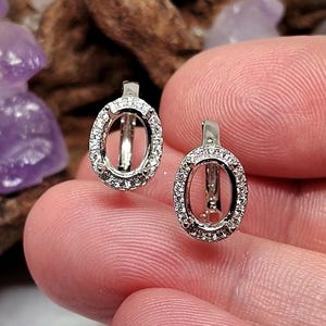 May include: A pair of silver-toned oval-shaped earring settings featuring a halo of small, clear, sparkling stones embedded around the perimeter. Each setting has a hinged clasp mechanism and an empty center designed to hold a gemstone.