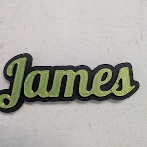 May include: A custom name sign featuring the name James in a cursive font. The letters are a metallic light green color with a textured surface, layered on top of a solid black backing that follows the outline of the script lettering.