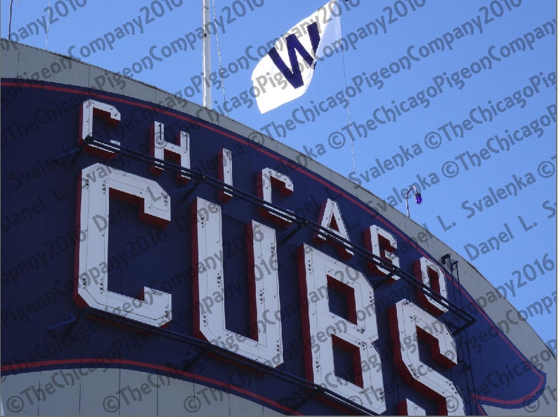 May include: A sign for the Chicago Cubs baseball team. The word "CUBS" is prominently displayed in large, white, illuminated letters against a navy blue background. A white flag with a blue "W" flies above. The sky is a clear blue.