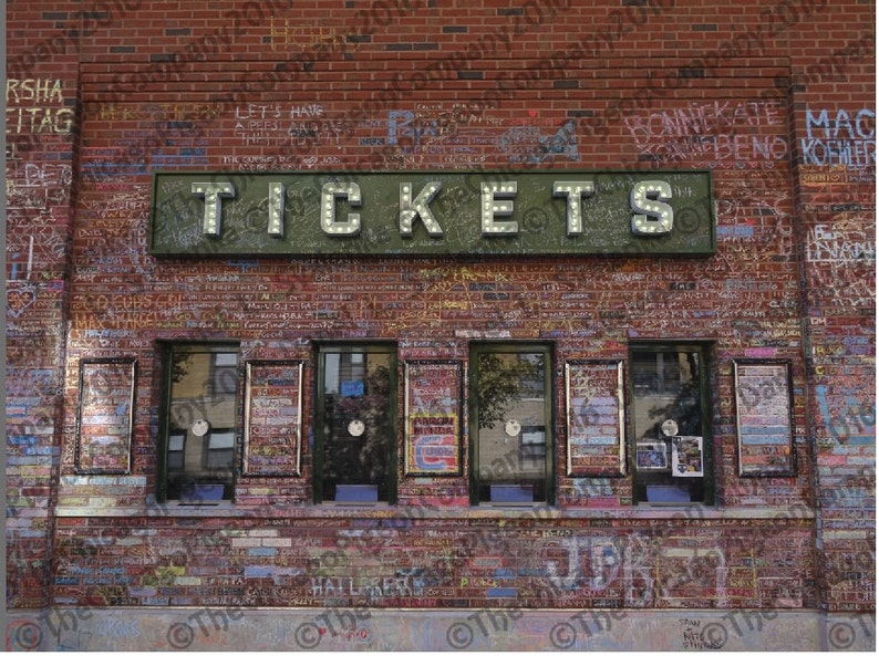 May include: A brick building with a "TICKETS" sign above five ticket windows. The sign has illuminated letters. The brick wall is covered in graffiti. The windows have posters and are framed in dark green.