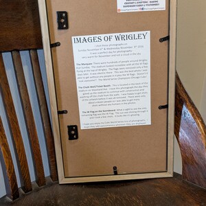 May include: Framed print titled "Images of Wrigley" with text about photographs taken at Wrigley Field. The frame is light brown, and the print includes text and a logo for The Chicago Pigeon Company.