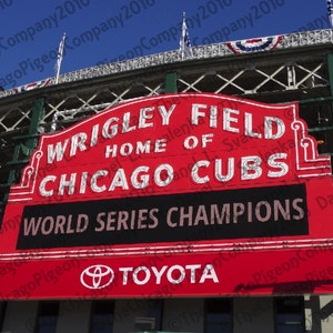 May include: A red sign at Wrigley Field, home of the Chicago Cubs, with the text "WRIGLEY FIELD HOME OF CHICAGO CUBS WORLD SERIES CHAMPIONS" and the Toyota logo. The sign is against a blue sky.