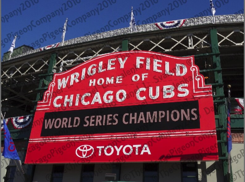 May include: A red sign at Wrigley Field, home of the Chicago Cubs, with the text "WRIGLEY FIELD HOME OF CHICAGO CUBS WORLD SERIES CHAMPIONS" and the Toyota logo. The sign is against a blue sky.