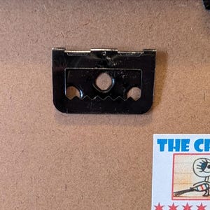 May include: Close-up of a black picture frame hanger with two holes and a sawtooth edge, along with a smaller metal piece. The background is a light brown surface. A small, colorful sticker is visible.