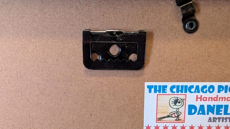 May include: Close-up of a black picture frame hanger with two holes and a sawtooth edge, along with a smaller metal piece. The background is a light brown surface. A small, colorful sticker is visible.
