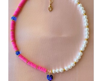 Puede incluir: Un collar vibrante con un diseño dividido. Una mitad está compuesta por cuentas de disco rosa brillante y detalles azules, mientras que la otra presenta perlas blancas. Un colgante de corazón azul cuelga del centro, con detalles metálicos dorados.