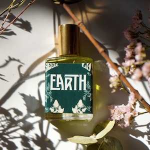 May include: A perfume bottle with a gold cap and a label that reads "EARTH" in white lettering. The label is dark green with white floral accents. The bottle is surrounded by dried flowers and foliage, casting shadows on a white surface.