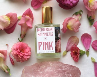 May include: A clear glass perfume bottle with a gold cap, labeled "PINK" by Wonderlust Botanicals, surrounded by pink flowers and a rose quartz crystal on a white surface. The bottle is labeled "100% NATURAL".