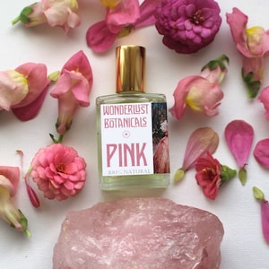 May include: A clear glass perfume bottle with a gold cap, labelled "PINK" by Wonderlust Botanicals, surrounded by pink flowers and a rose quartz crystal on a white surface. The bottle is labelled "100% NATURAL".