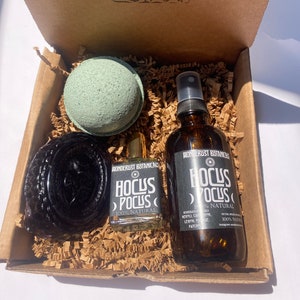 May include: A cardboard gift box containing a light green spherical bath bomb, a dark textured soap bar, a small glass bottle, and a larger amber glass spray bottle. Both bottles feature black labels with the text Wonderlust Botanicals Hocus Pocus.