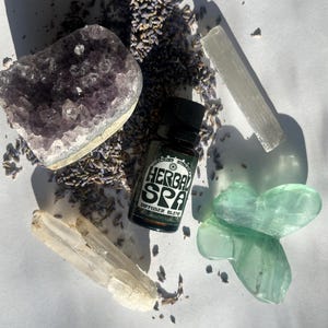 Peut inclure: Composition de produits naturels : une géode d'améthyste violette, de la lavande séchée, une bouteille de mélange pour diffuseur "Herbal Spa" et des cristaux blancs et verts. Les articles sont disposés sur une surface blanche.