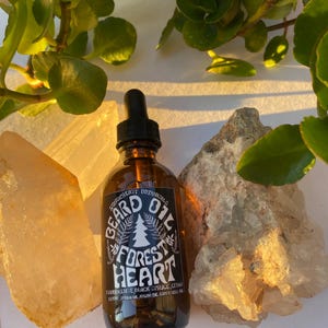 Peut inclure: Une bouteille en verre brun d'huile à barbe avec un compte-gouttes noir, étiquetée "Forest Heart". L'étiquette présente un graphisme d'arbre. La bouteille est entourée de cristaux et de verdure, suggérant un produit naturel.