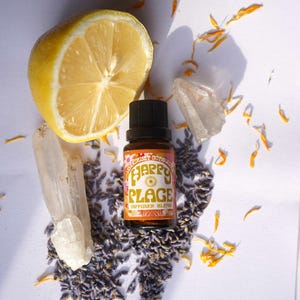 May include: A still life arrangement with a halved lemon, a small amber glass bottle labelled "Happy Place Diffuser Blend", quartz crystals, lavender buds, and orange flower petals. The bottle has a black cap and a colourful label.