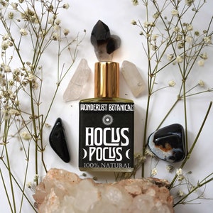 Peut inclure: Un flacon de parfum avec un bouchon doré et une étiquette noire indiquant "Hocus Pocus 100% Natural". Le flacon est entouré de cristaux, de pierres et de brins de fleurs blanches. La marque "Wonderlust Botanicals" est également visible.